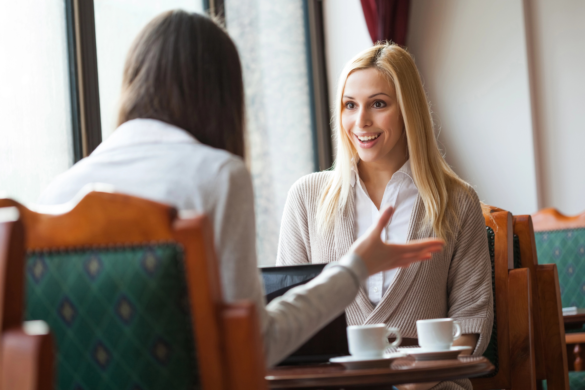 Eine blonde Frau in einem Business-Casual Outfit diskutiert lebhaft mit einer Vertrauens-Beraterin in einem Café.